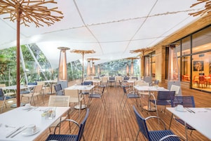 Zona pranzo all'aperto con tavoli e sedie in legno, ampie finestre e vista sul verde.