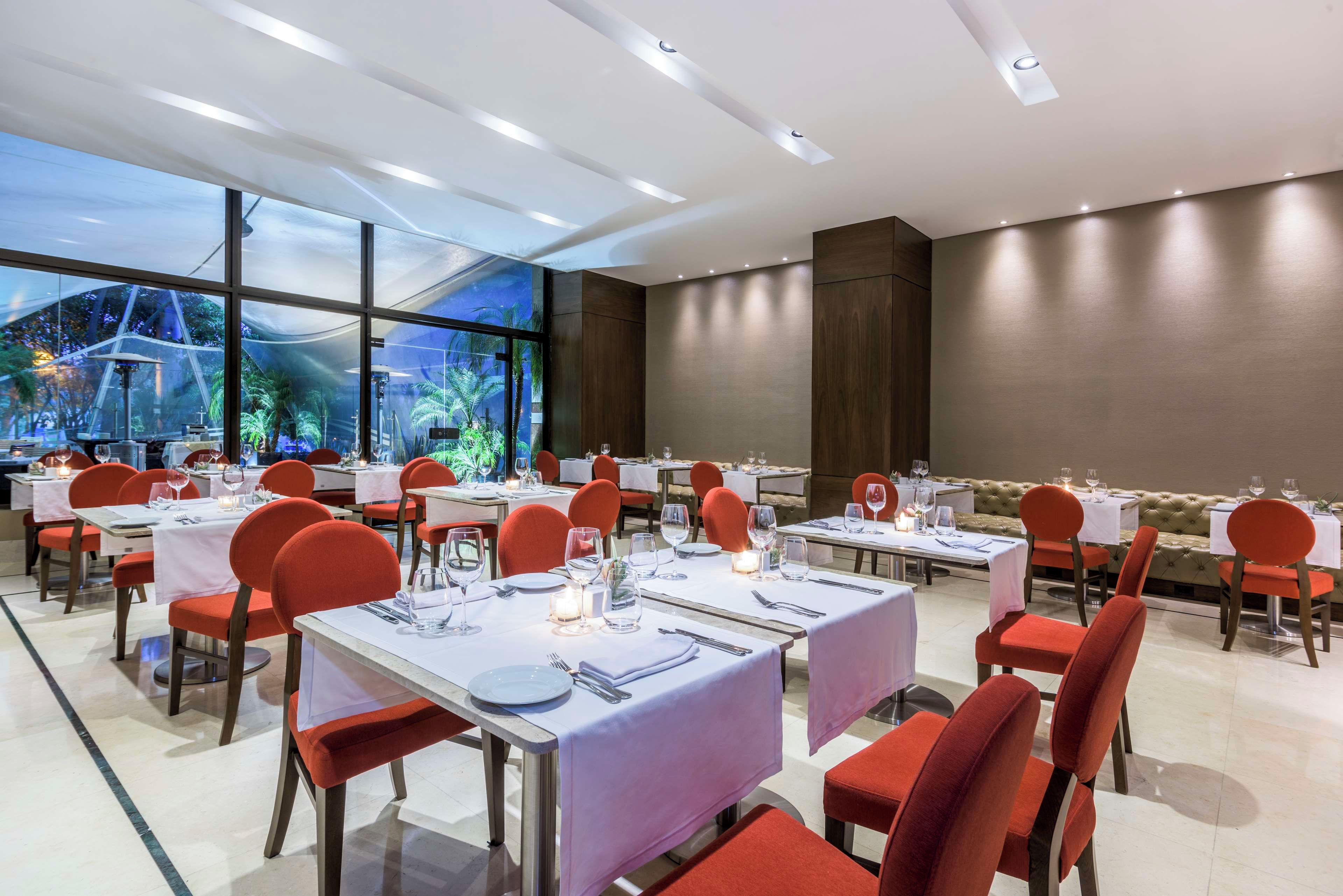 A restaurant with tables set for dining, red chairs, and a view of greenery through large windows.