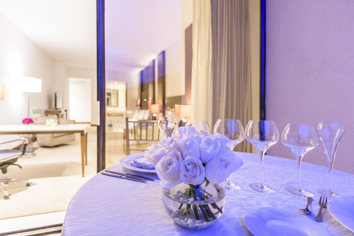 A hotel dining area with a round table set for a meal, featuring a clear glass bowl of white roses and wine glasses.
