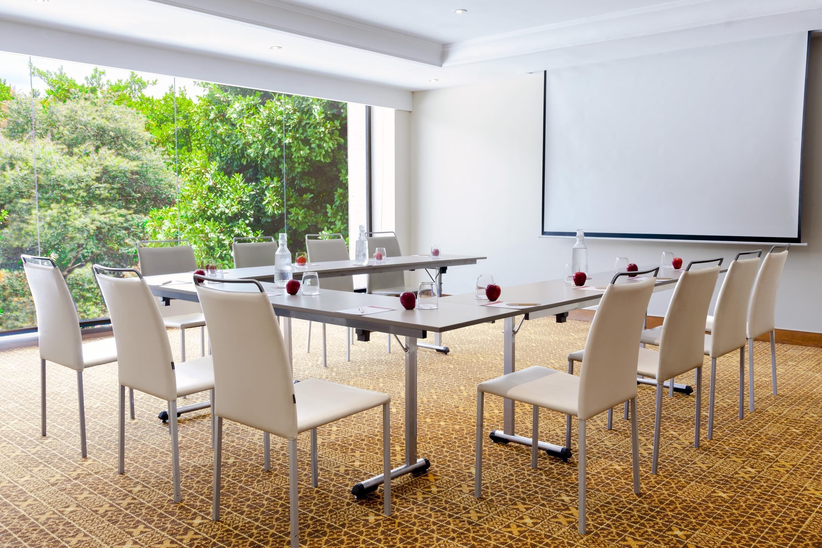 Un salón de conferencias con una amplia mesa, sillas, una pantalla de proyector y vistas al exterior con vegetación.
