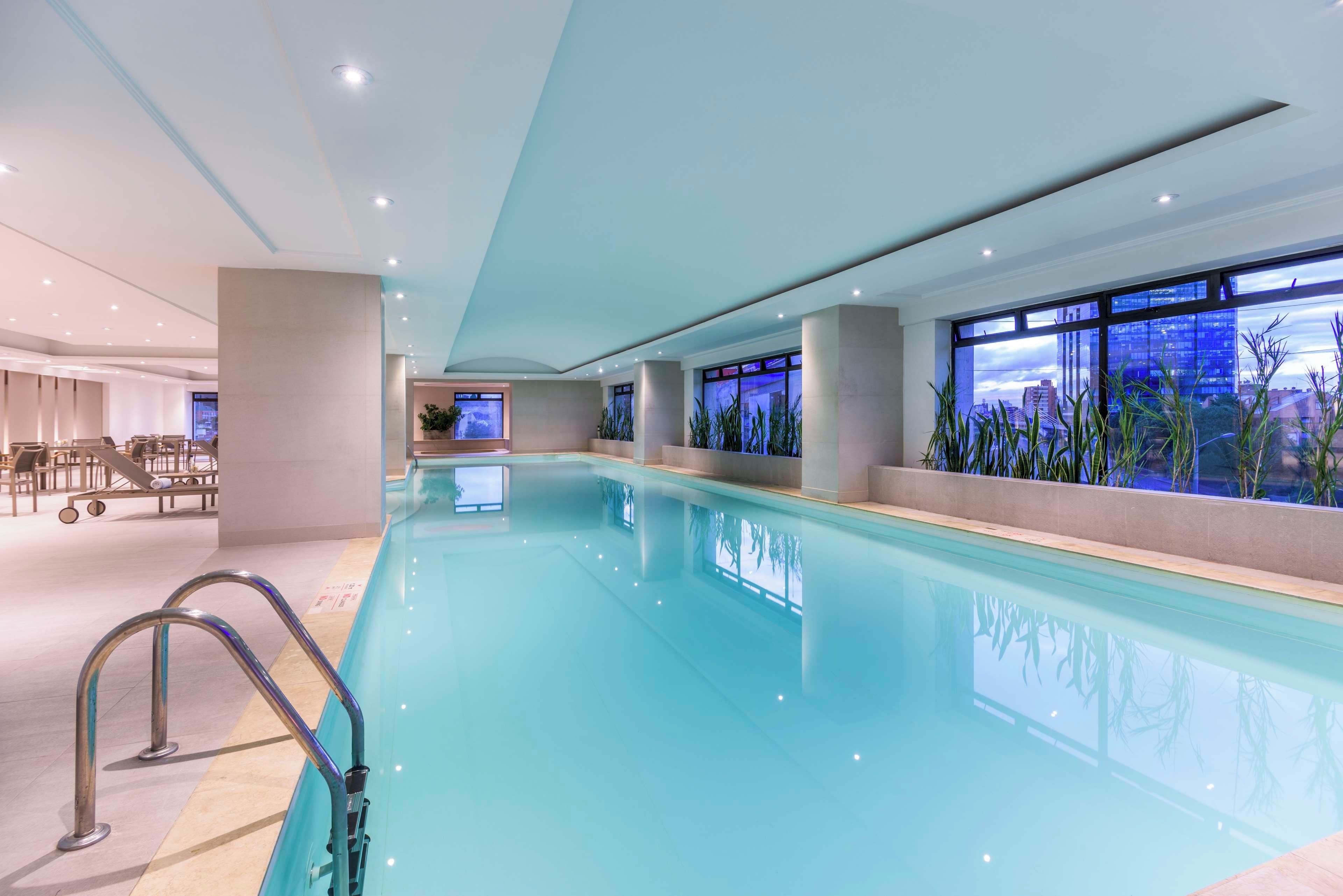 A large indoor swimming pool with clear water, surrounded by seating areas and large windows offering views of the cityscape.