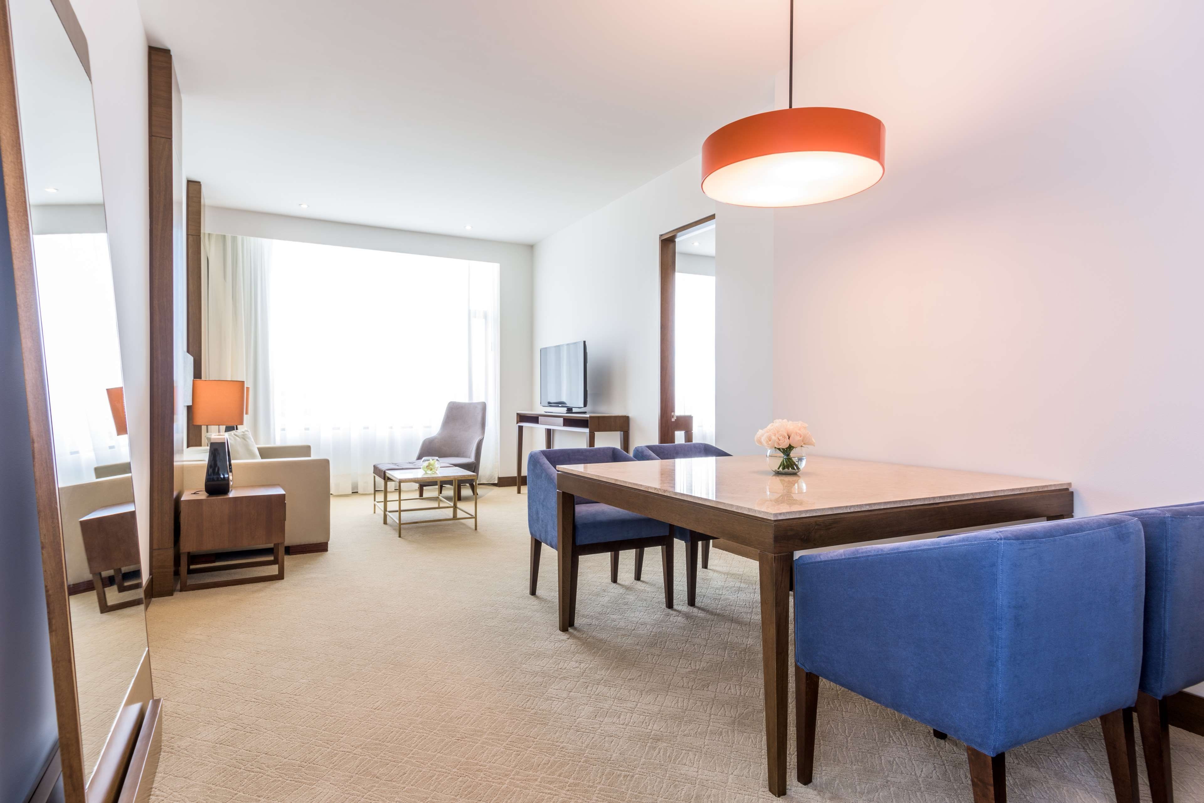 Habitación de hotel con zona de comedor, sofá, televisor y una ventana grande.
