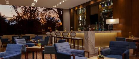 A modern bar area with blue upholstered chairs, wooden tables, and a bar stocked with bottles.