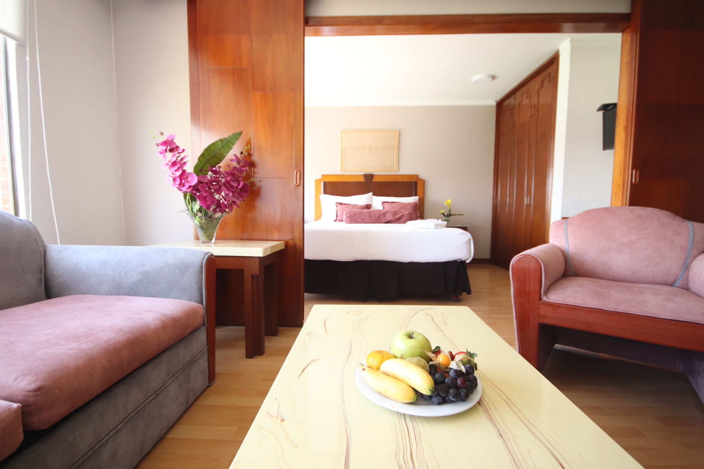Quarto de hotel com cama, área de estar com mesa e cadeiras, e uma fruteira sobre a mesa.