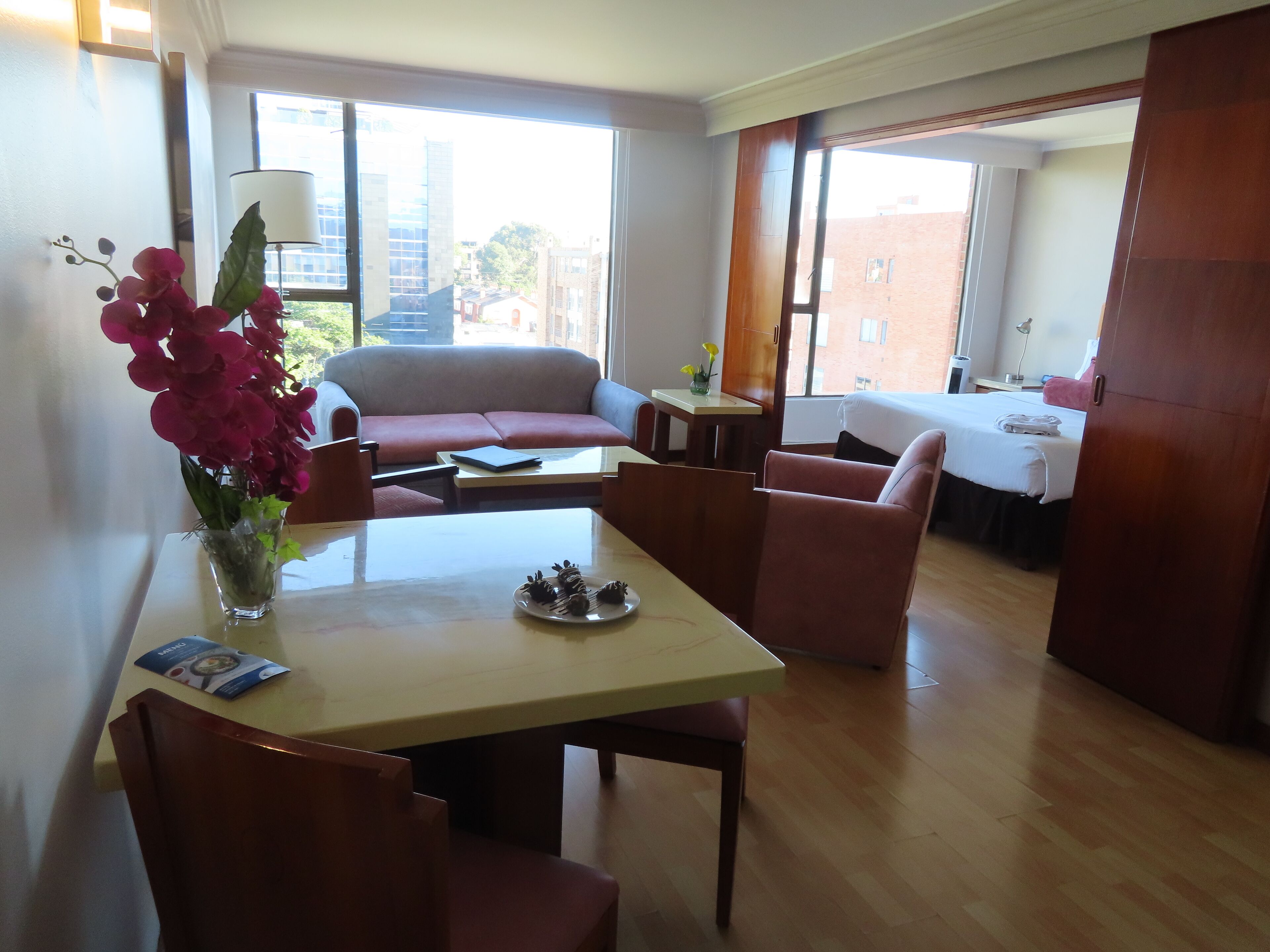 Una sala moderna con un sofá, una mesa de comedor con sillas, un jarrón de flores y vistas a la ciudad a través de amplios ventanales.
