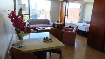 Una sala moderna con un sofá, una mesa de comedor con sillas, un jarrón de flores y vistas a la ciudad a través de amplios ventanales.