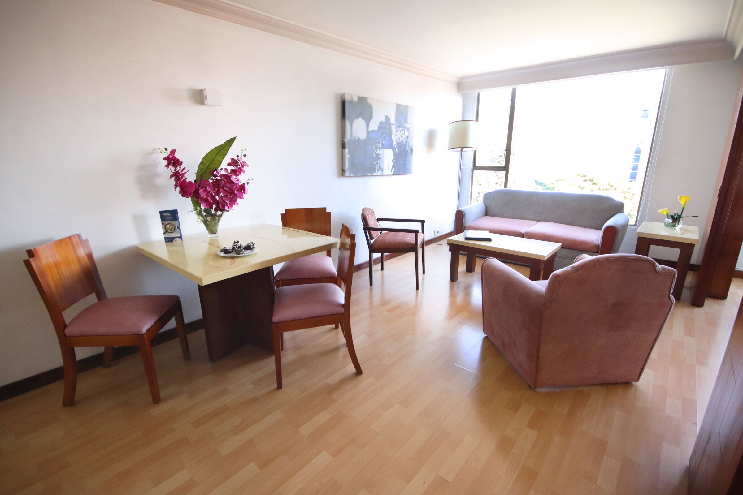 A dining area with a table, chairs, and a vase of flowers. There is a sofa and a lamp in the background.