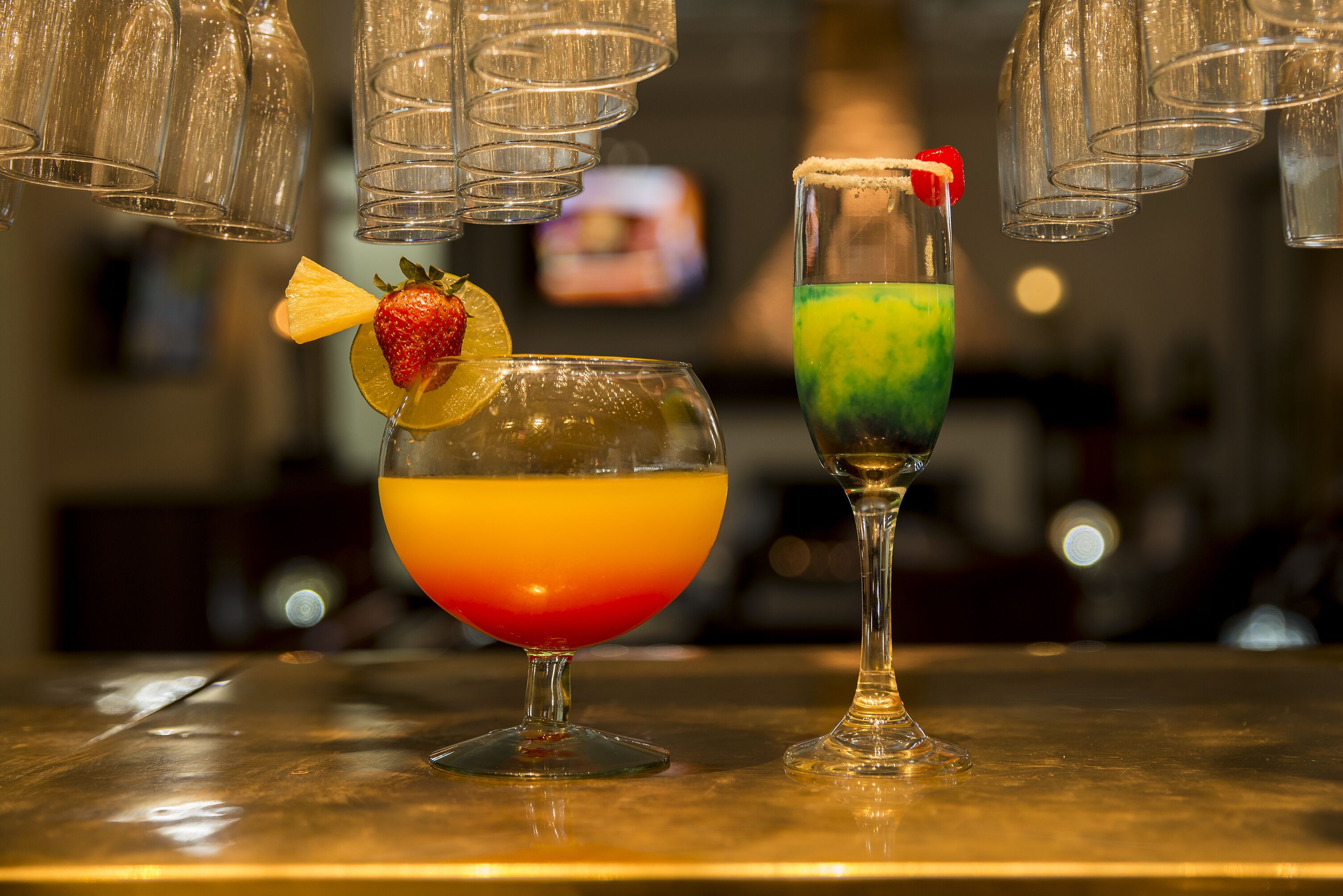 Dois cocktails sobre um balcão de bar, com copos pendurados acima.