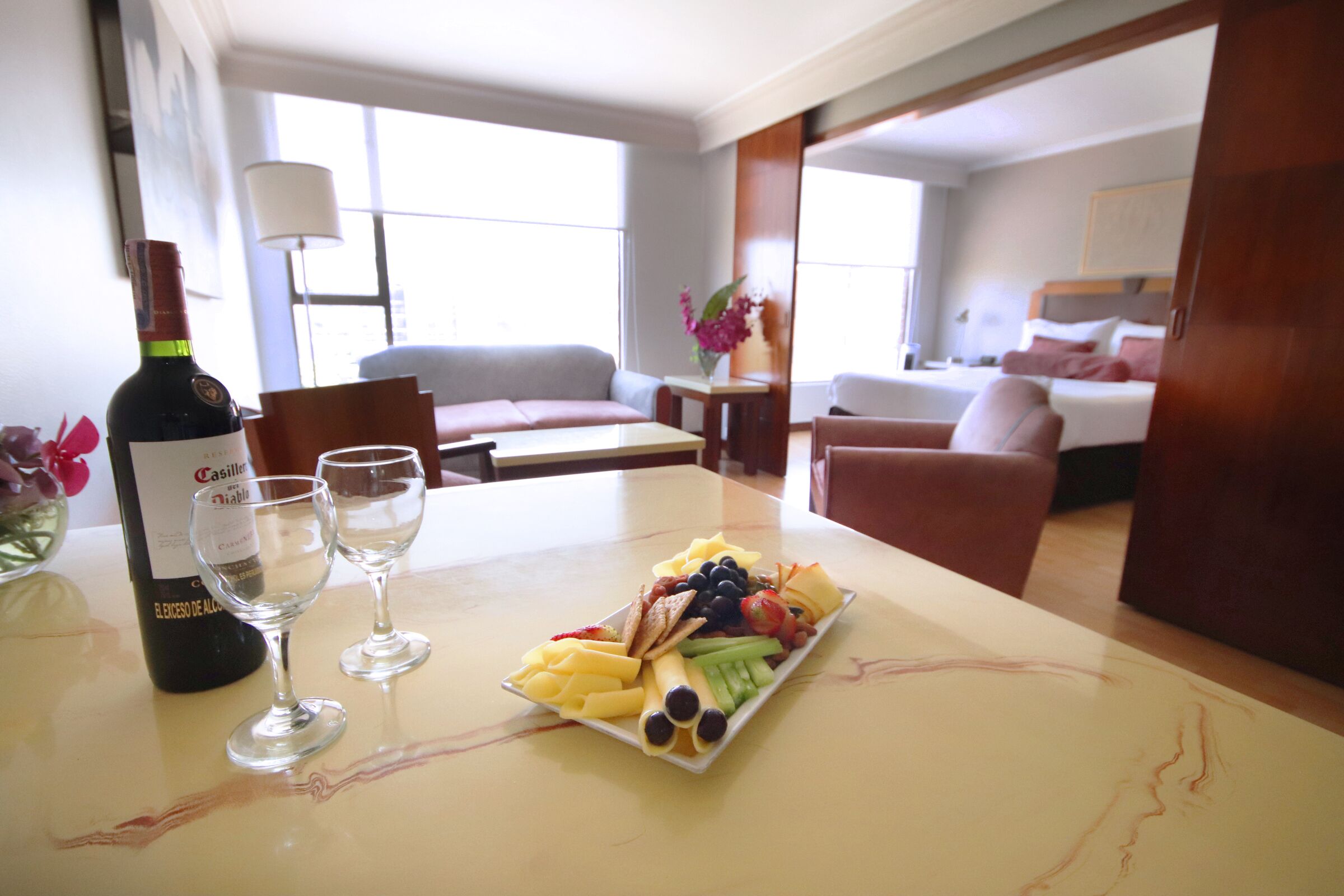 A hotel room with a dining table set with a bottle of wine, two glasses, and a fruit platter.