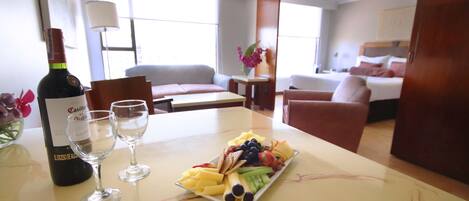 A hotel room with a dining table set with a bottle of wine, two glasses, and a fruit platter.