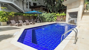 Une piscine au fond recouvert de carreaux bleus, entourée de chaises longues et agrémentée d’une cascade.