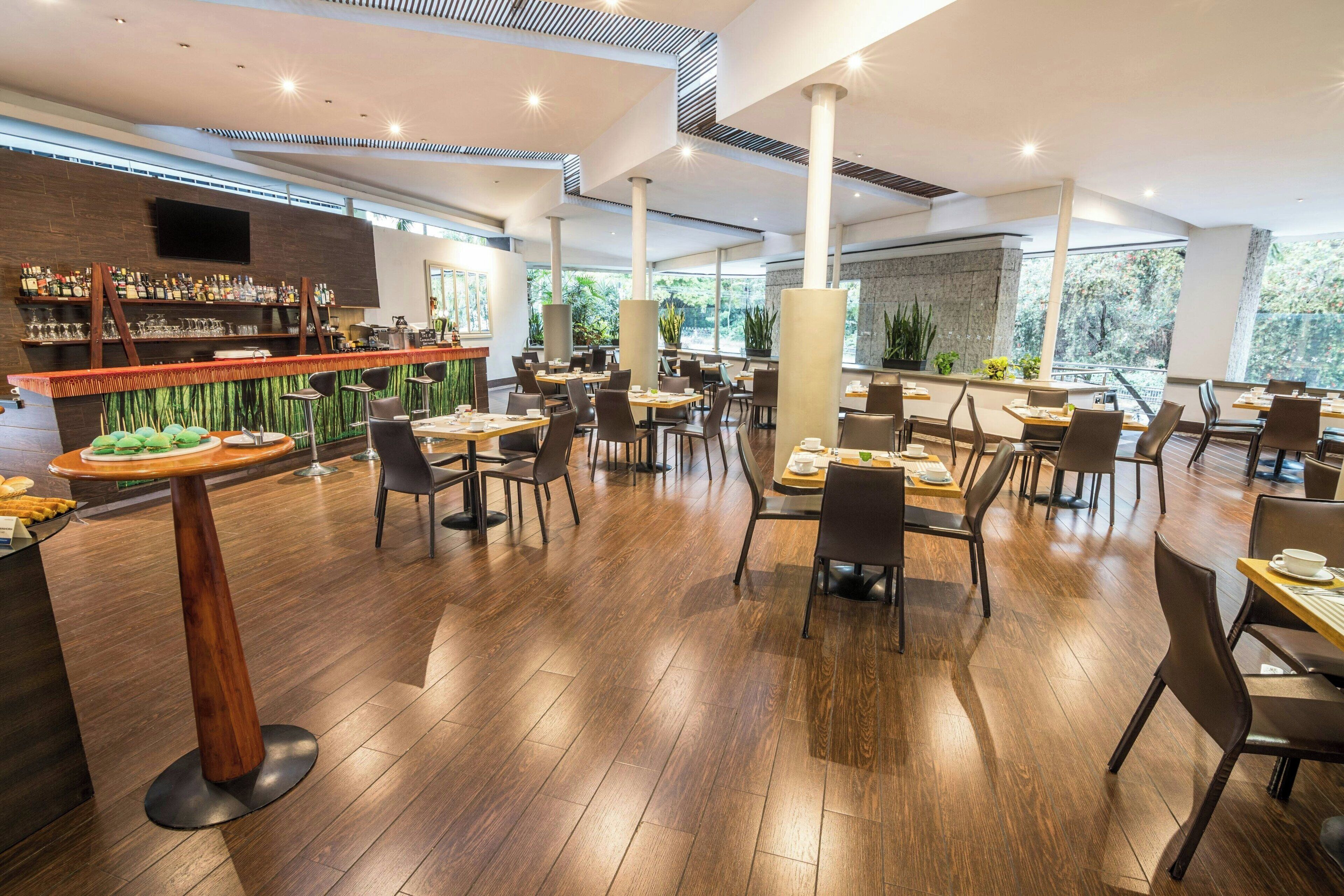 Um restaurante moderno com piso de madeira, pé direito alto e área de bar com diversas bebidas e petiscos.