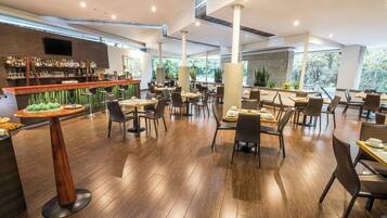 Um restaurante moderno com piso de madeira, pé direito alto e área de bar com diversas bebidas e petiscos.