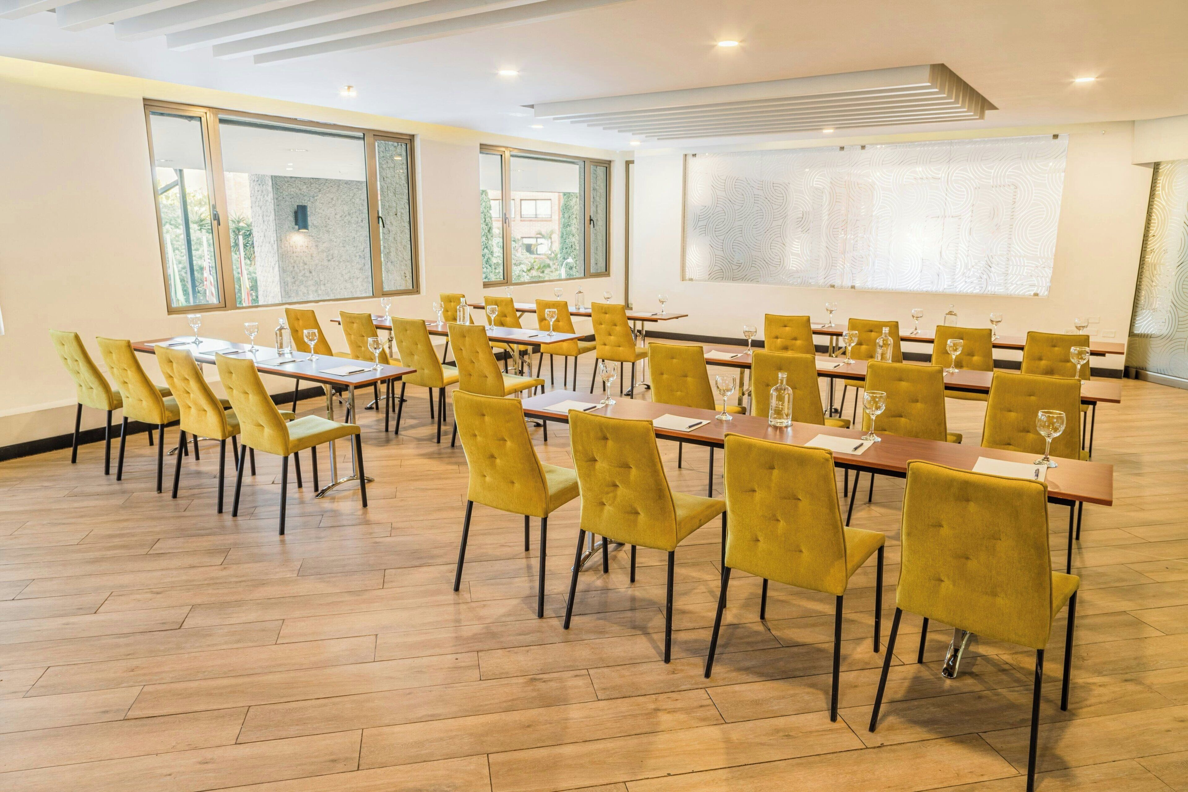 A spacious dining area with multiple tables and yellow chairs, large windows, and a modern ceiling design.