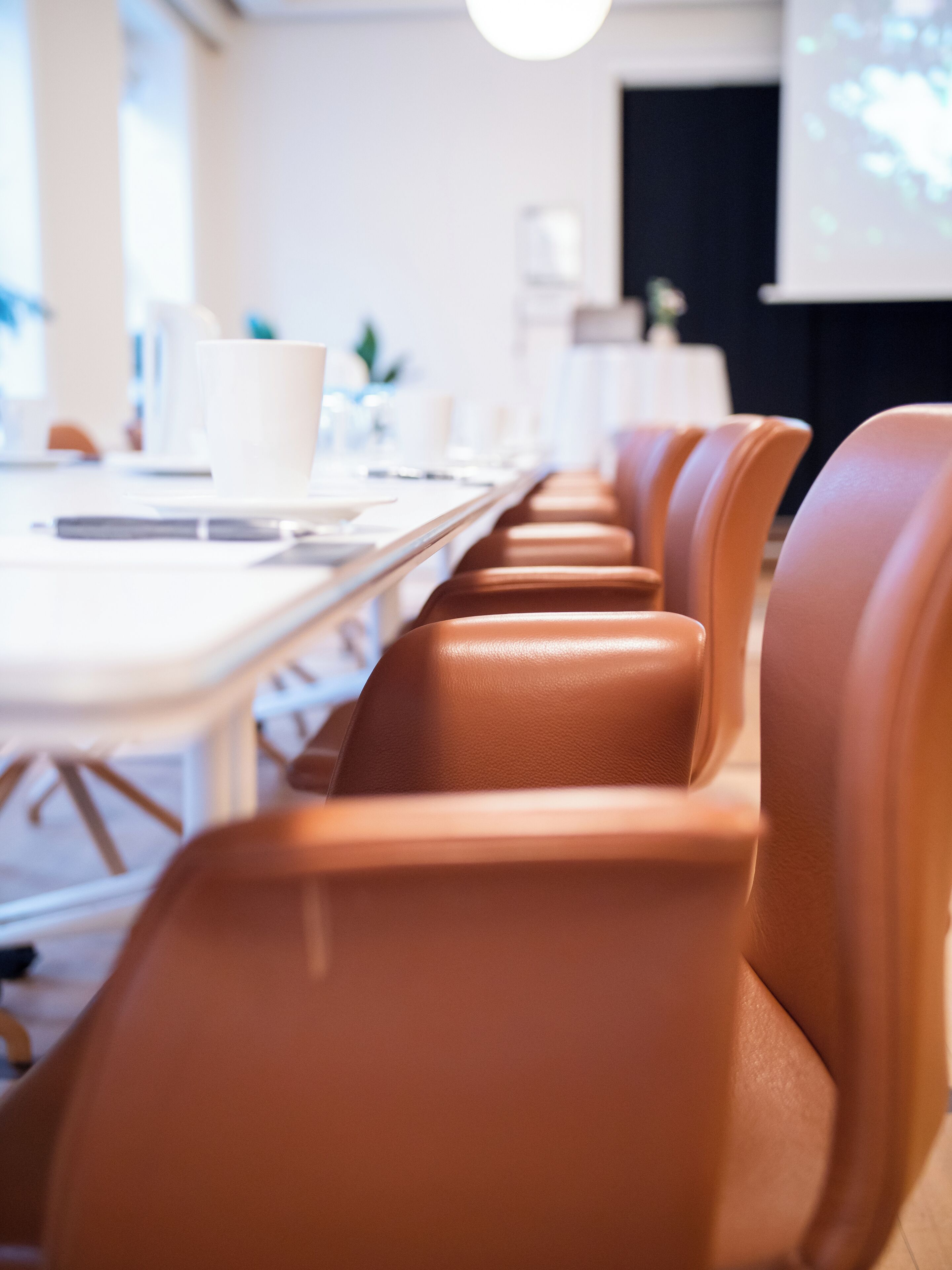 Un salón de conferencias con sillas de cuero y una mesa larga, equipado con computadoras portátiles y una pantalla de proyector.