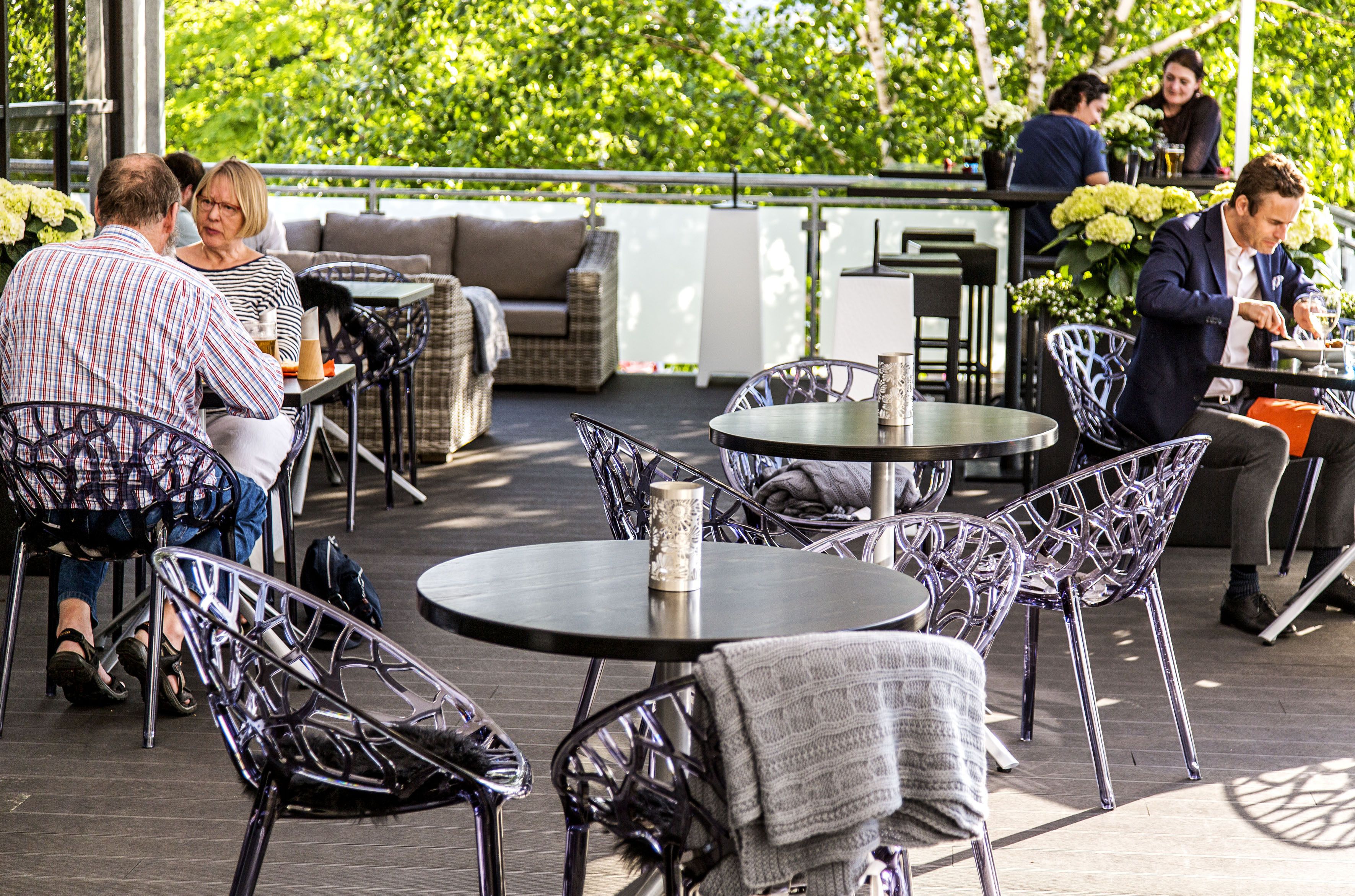 An outdoor patio with round tables and wicker chairs, surrounded by greenery.