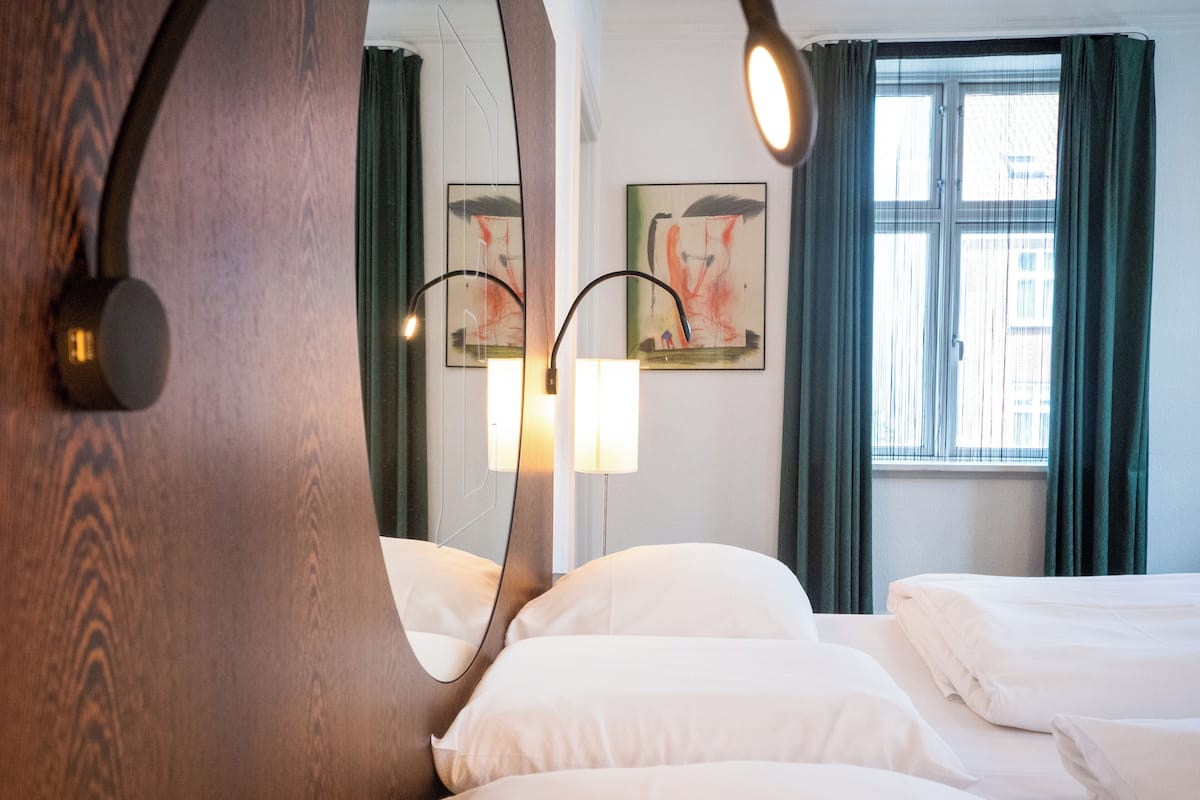 Quarto de hotel com um espelho grande, duas camas, uma janela com cortinas e obras de arte na parede.