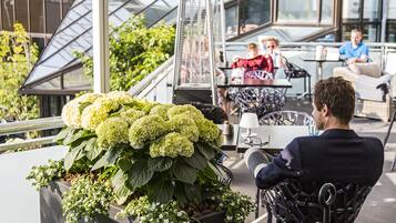 Una terraza en la azotea con mobiliario, una maceta con hortensias y un área acristalada donde personas están cenando.