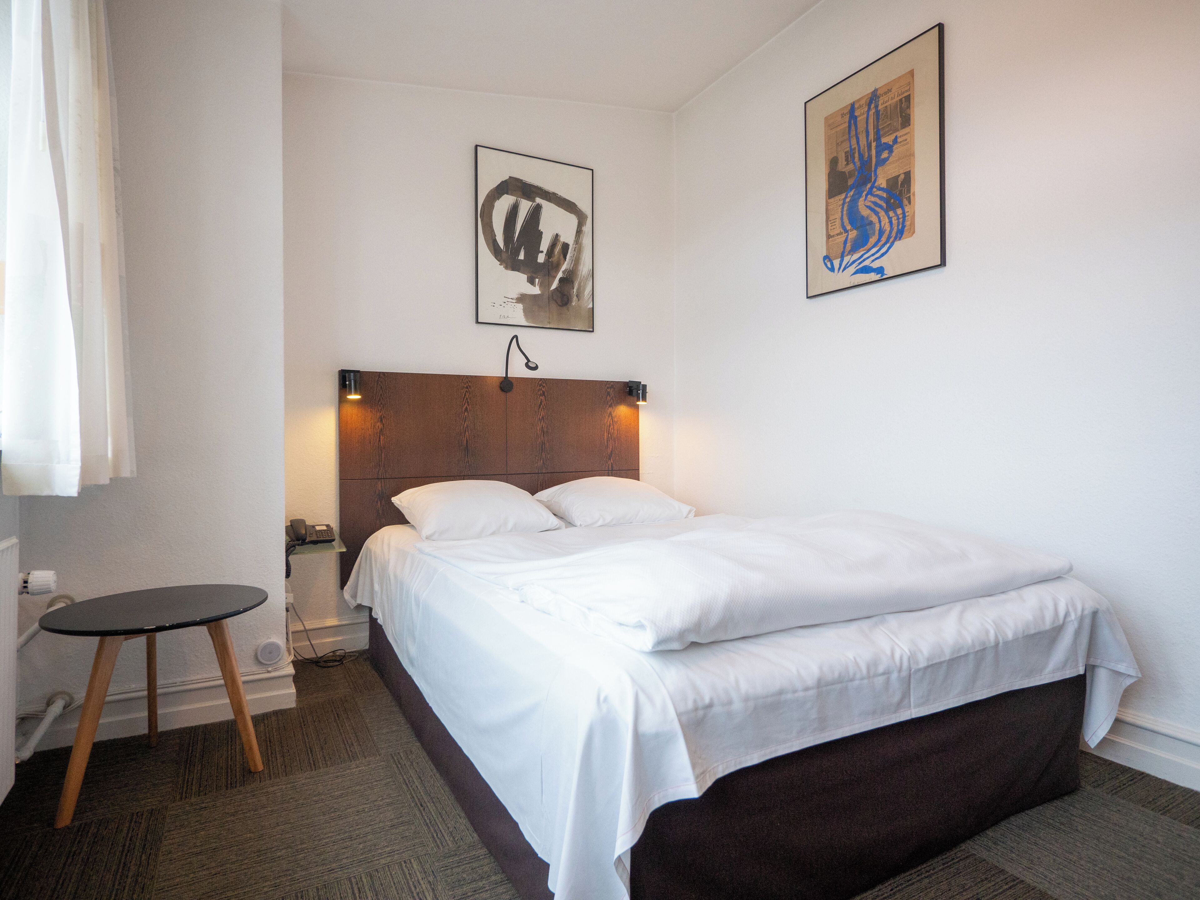 A hotel room with a wooden headboard, a single bed, a small round table, and two wall-mounted artworks.