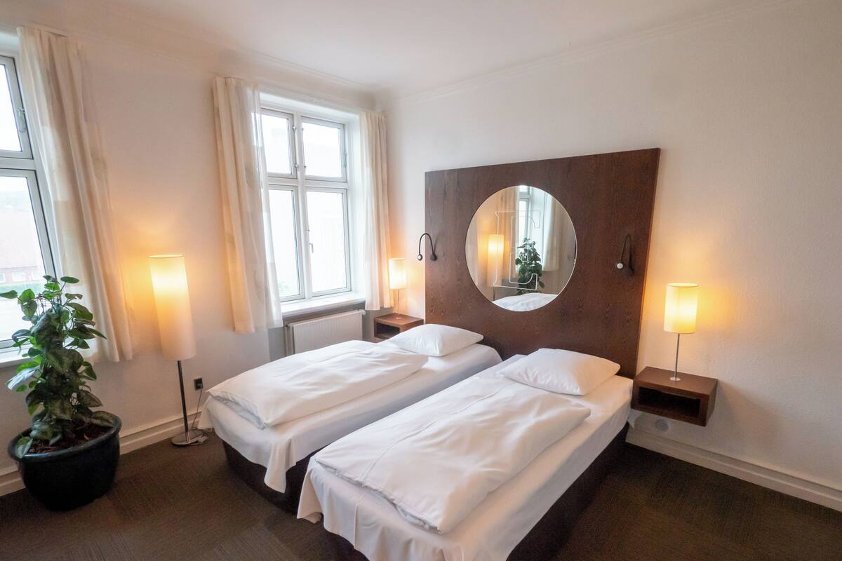 Quarto de hotel com duas camas de solteiro, um espelho grande, abajures, um vaso de plantas e uma janela com cortinas.