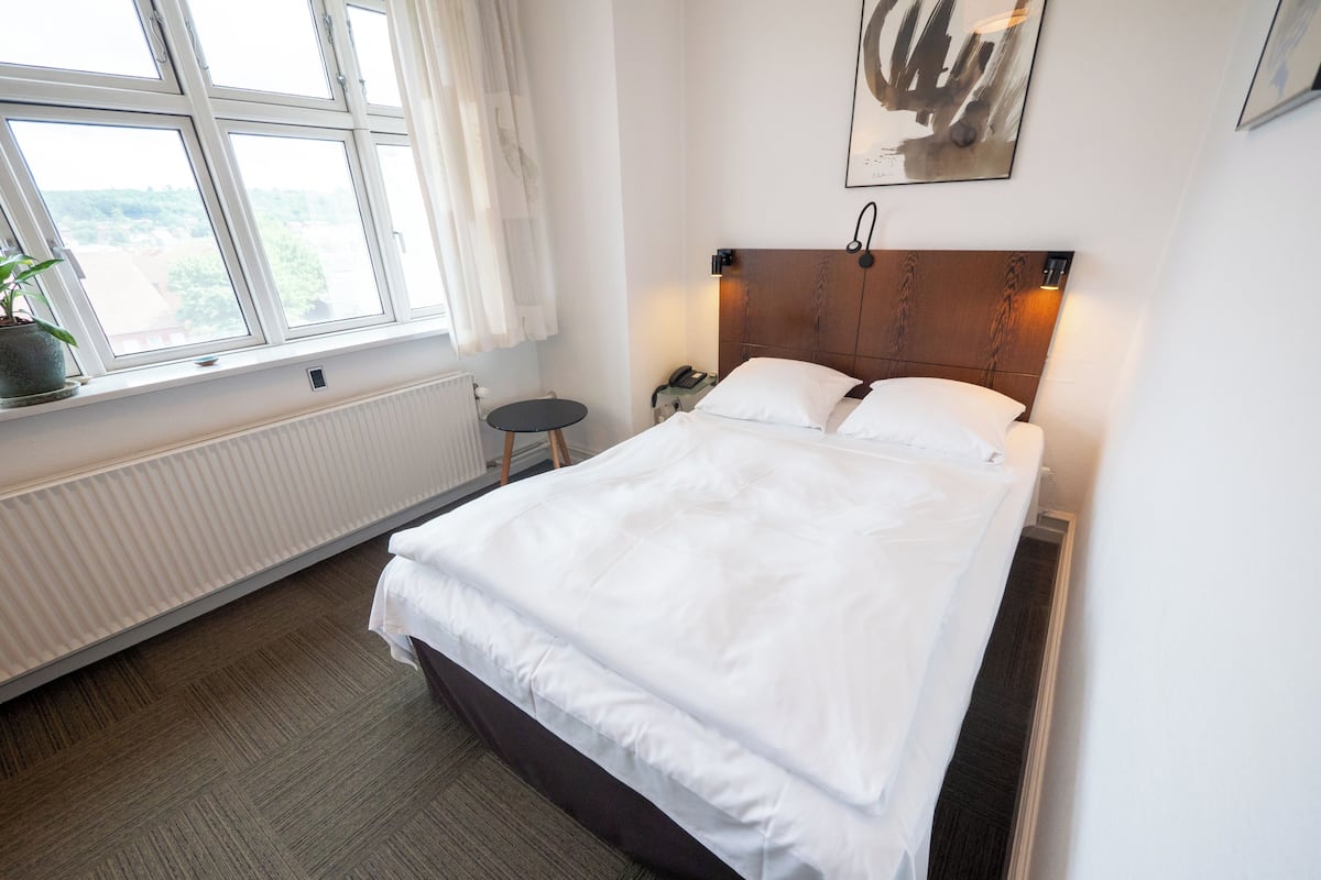 Cama bem arrumada num quarto de hotel com vista para o exterior através de uma janela, uma mesa de cabeceira e um telefone fixado na parede.