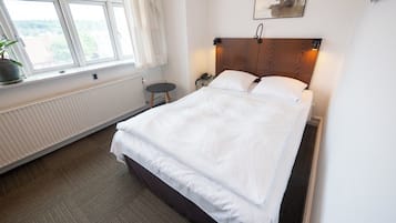 A neatly made bed in a hotel room with a view of the outdoors through a window, a bedside table, and a wall-mounted telephone.