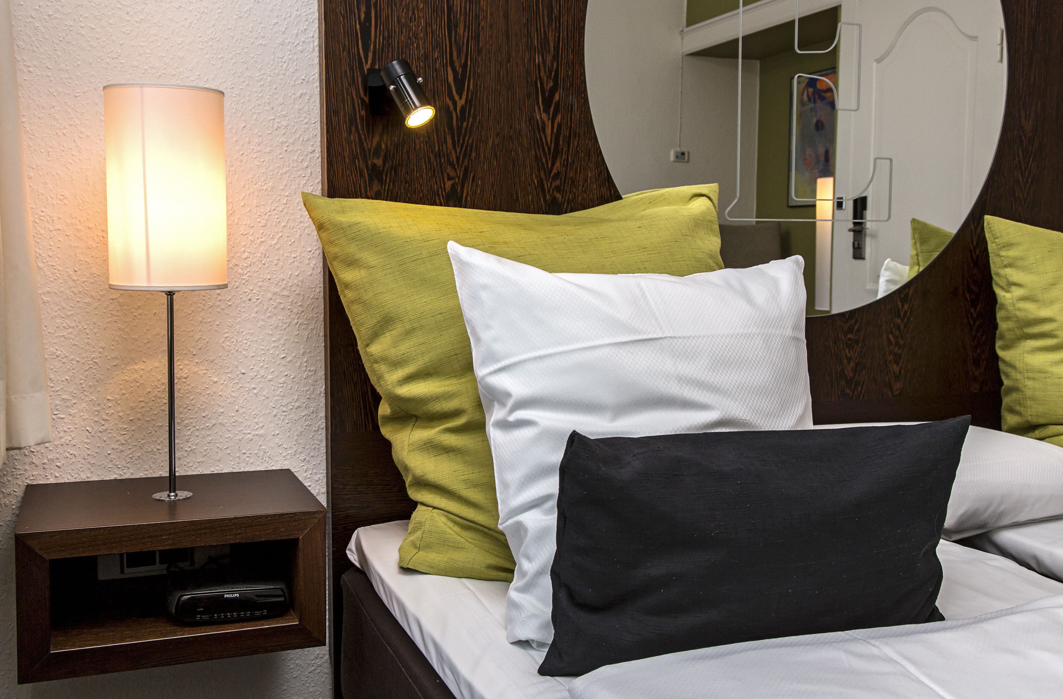 A neatly made bed with white and green pillows, a bedside lamp, and a small wooden nightstand with a remote control.