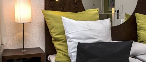 A neatly made bed with white and green pillows, a bedside lamp, and a small wooden nightstand with a remote control.