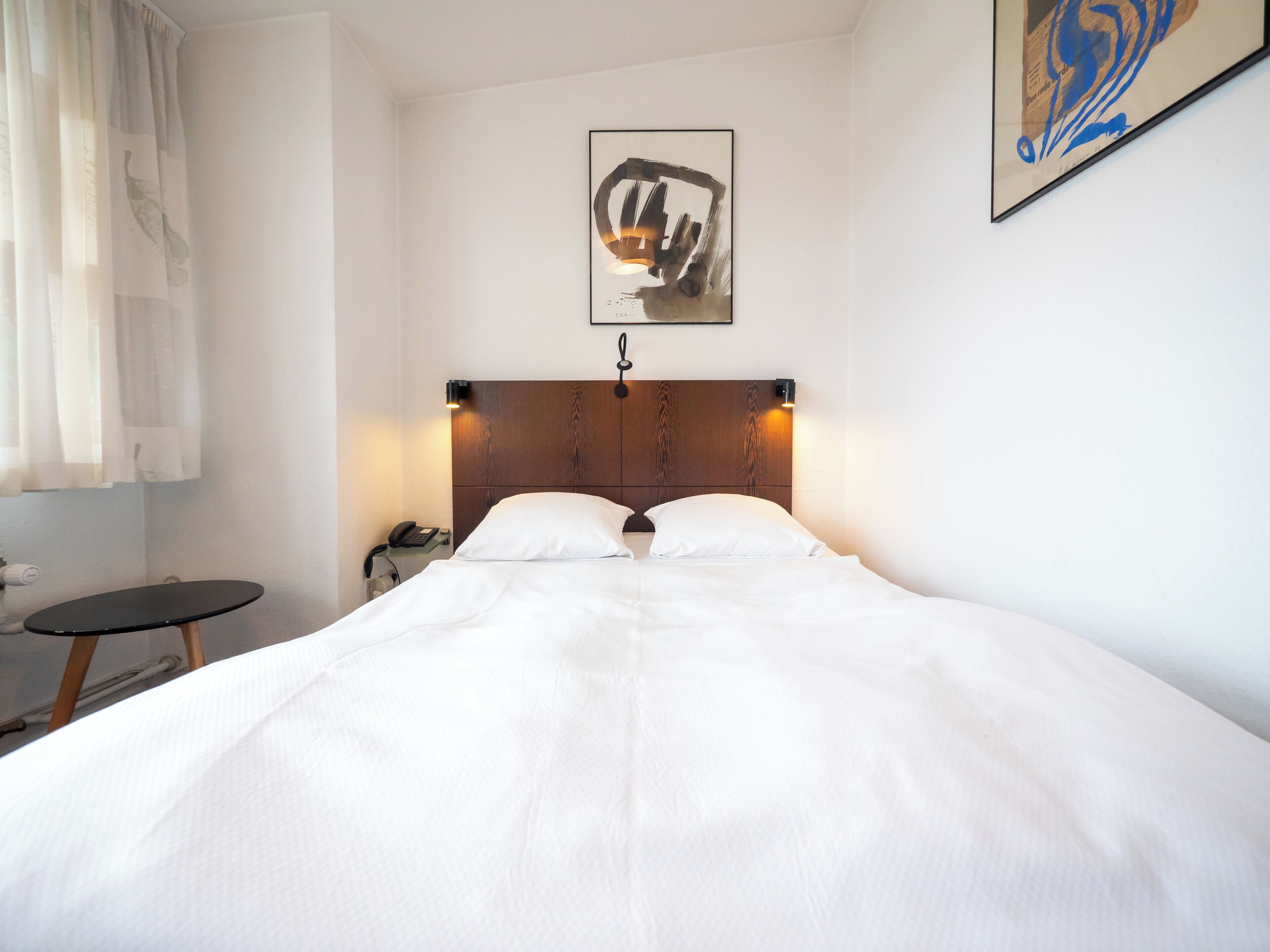 Un lit bien fait dans une chambre d’hôtel, avec une tête de lit en bois, une table de chevet avec une lampe et un tableau abstrait encadré accroché au mur.