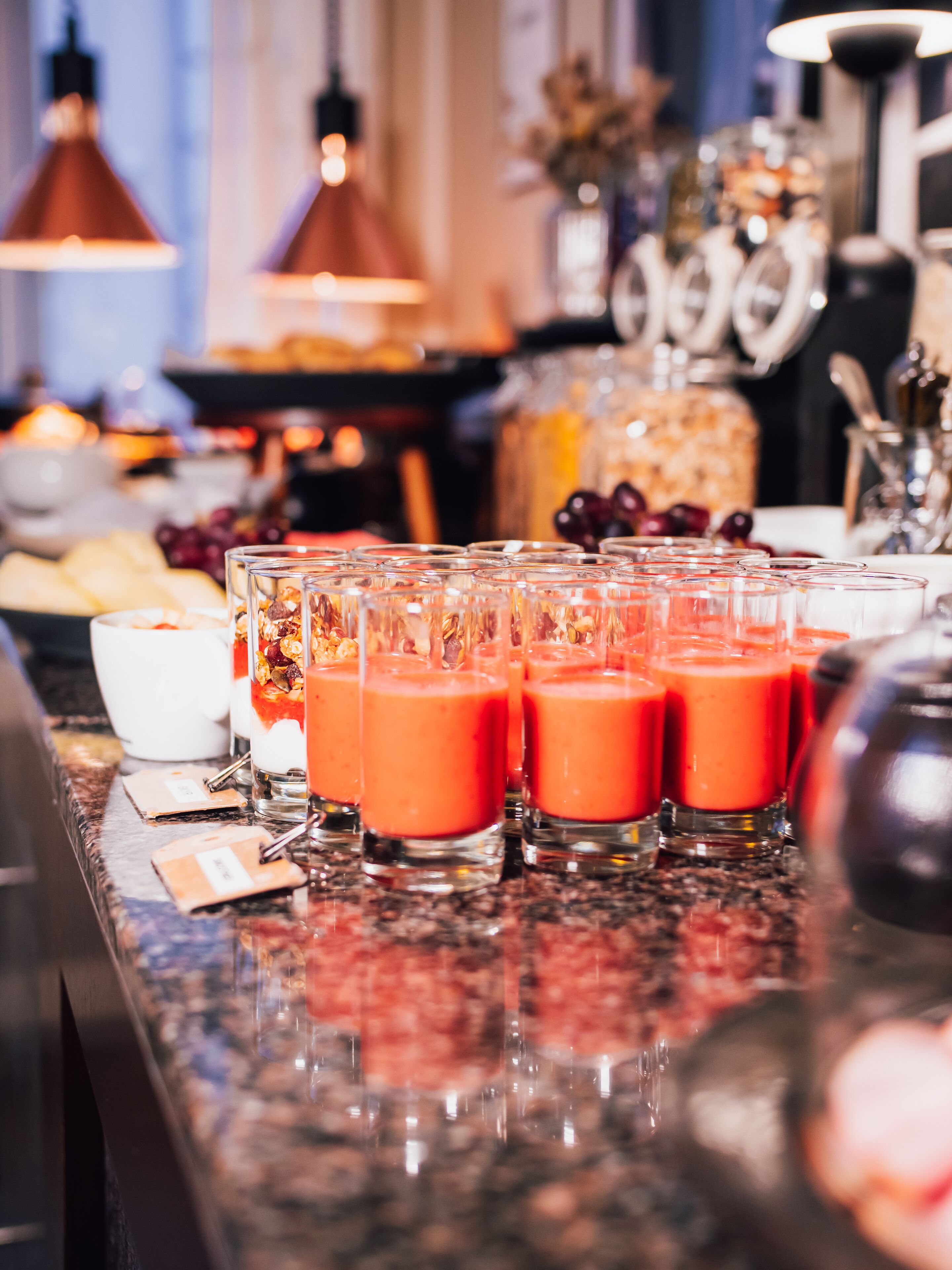 Una zona de bar con varios vasos de una bebida roja, una jarra blanca y diferentes frutas y aperitivos sobre la barra.