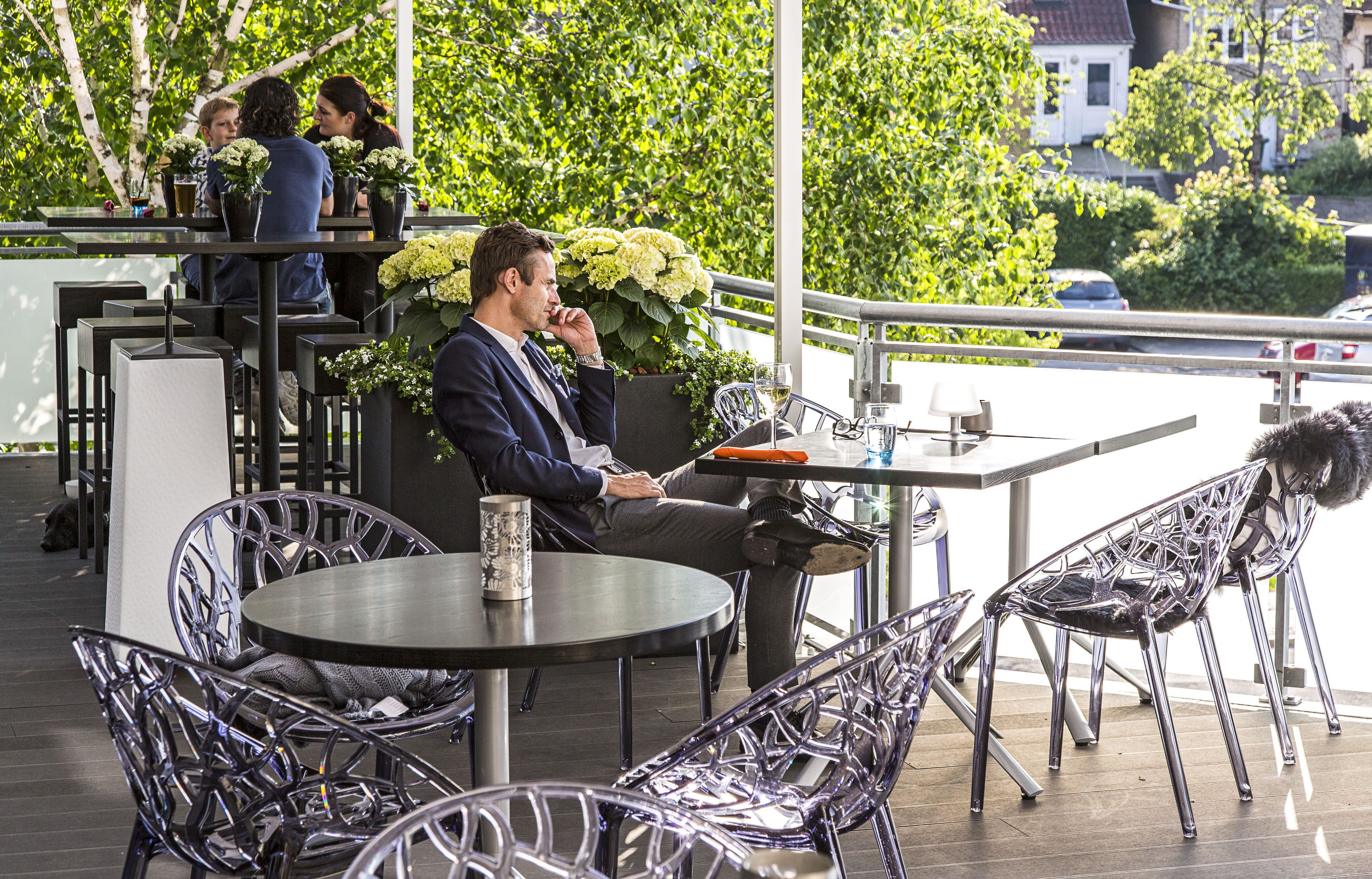 An outdoor café with round tables and metal chairs, surrounded by greenery and a railing.