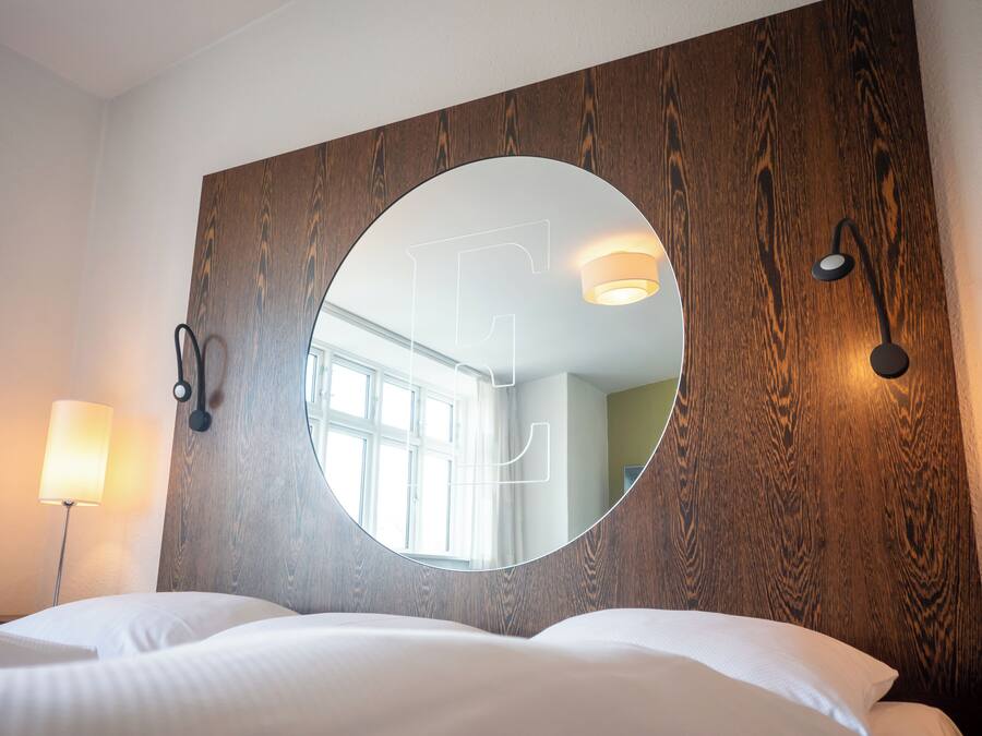 A hotel room with a large round mirror, wooden headboard, bedside lamp, and a bed with white linens.