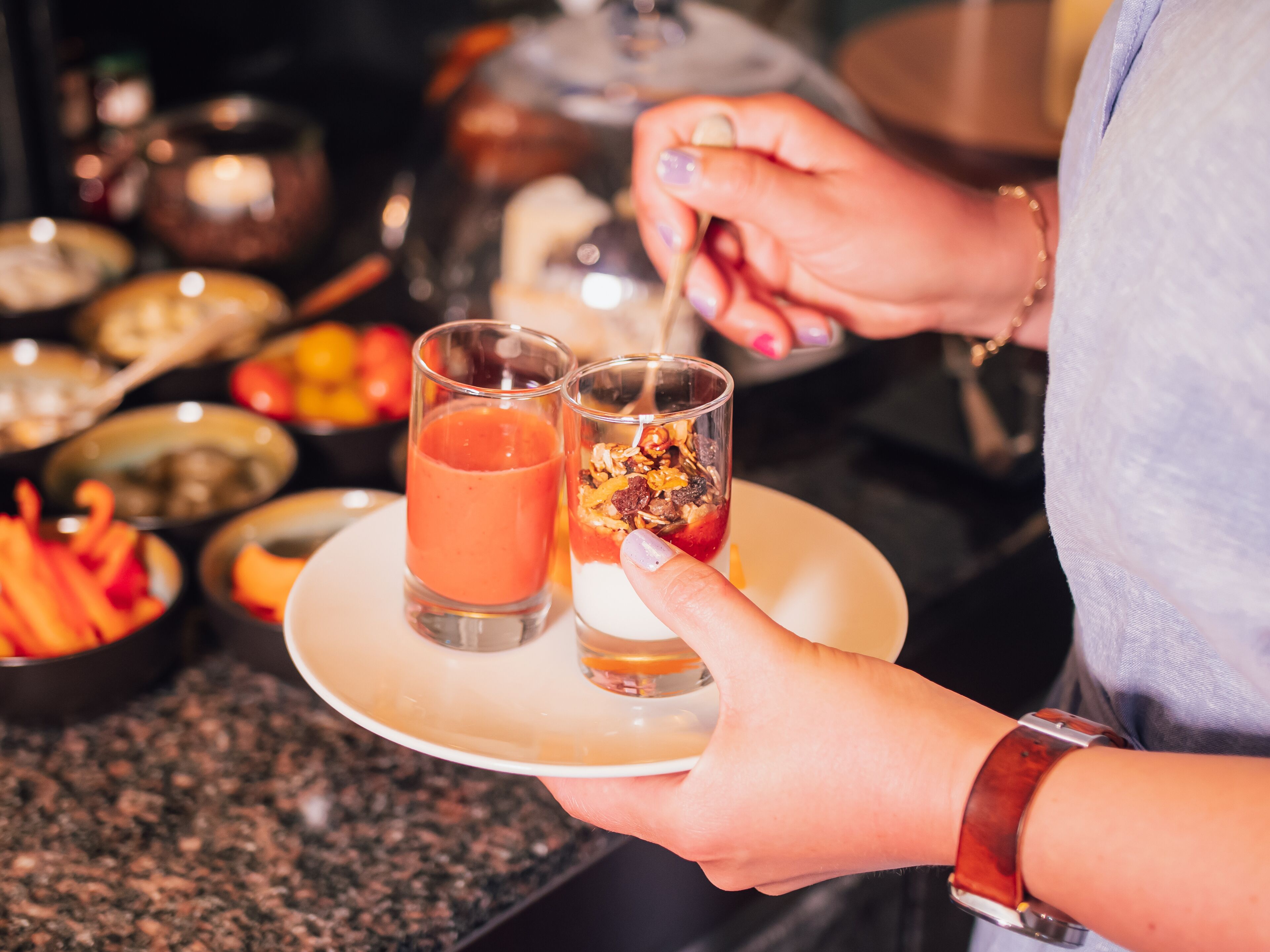 A person holding a plate with two drinks and a dessert, with various food items in the background.