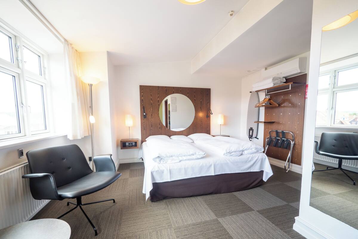Quarto de hotel com uma cama, uma escrivaninha, uma cadeira, um espelho, um abajur e uma janela.
