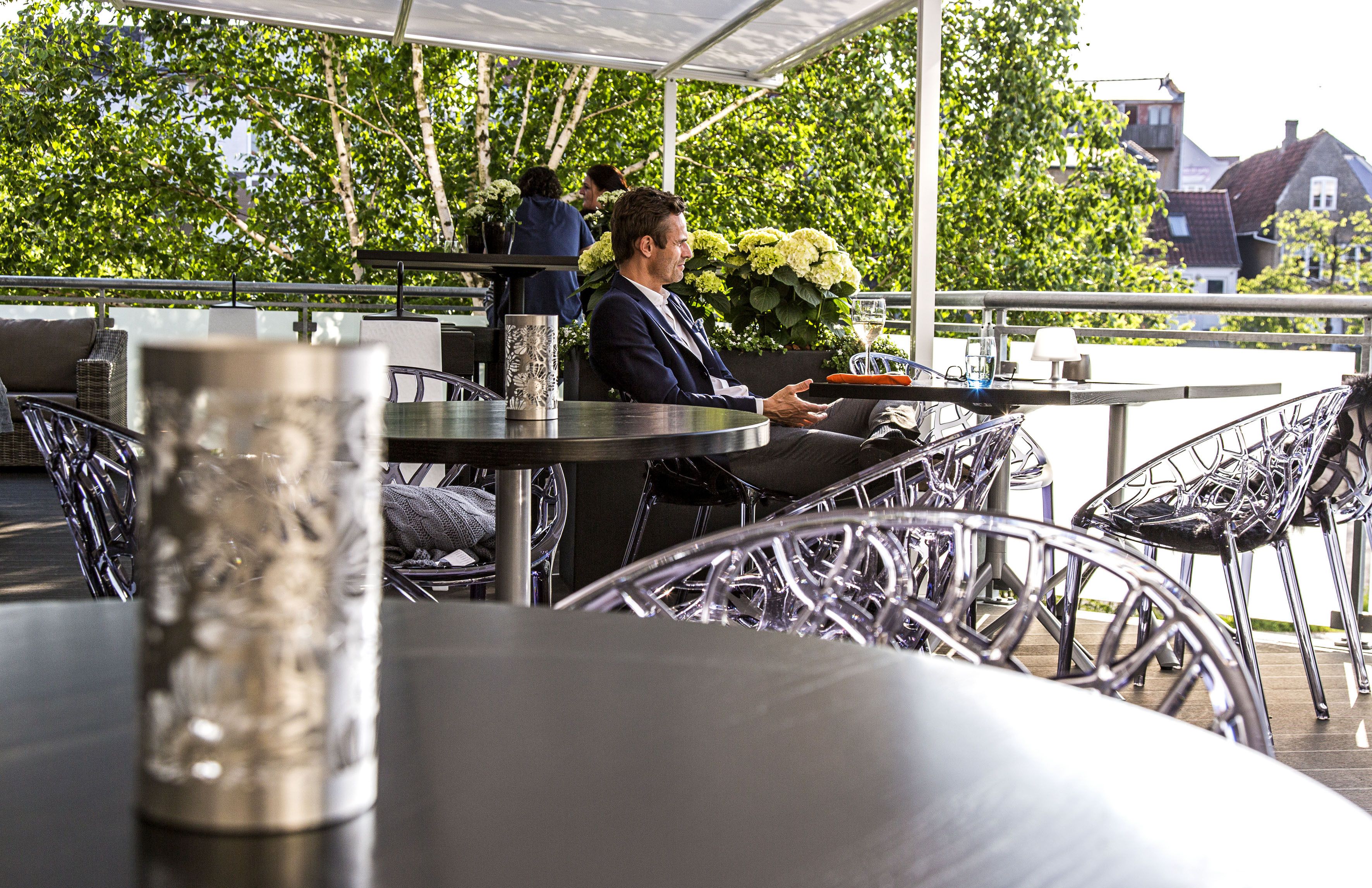 A covered outdoor seating area with metal chairs and tables, a potted plant, and a person seated at a table.