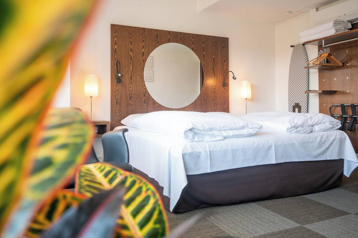 Quarto de hotel com uma cama grande, cabeceira de madeira, espelho redondo, abajures e um guarda-roupa com roupas penduradas.
