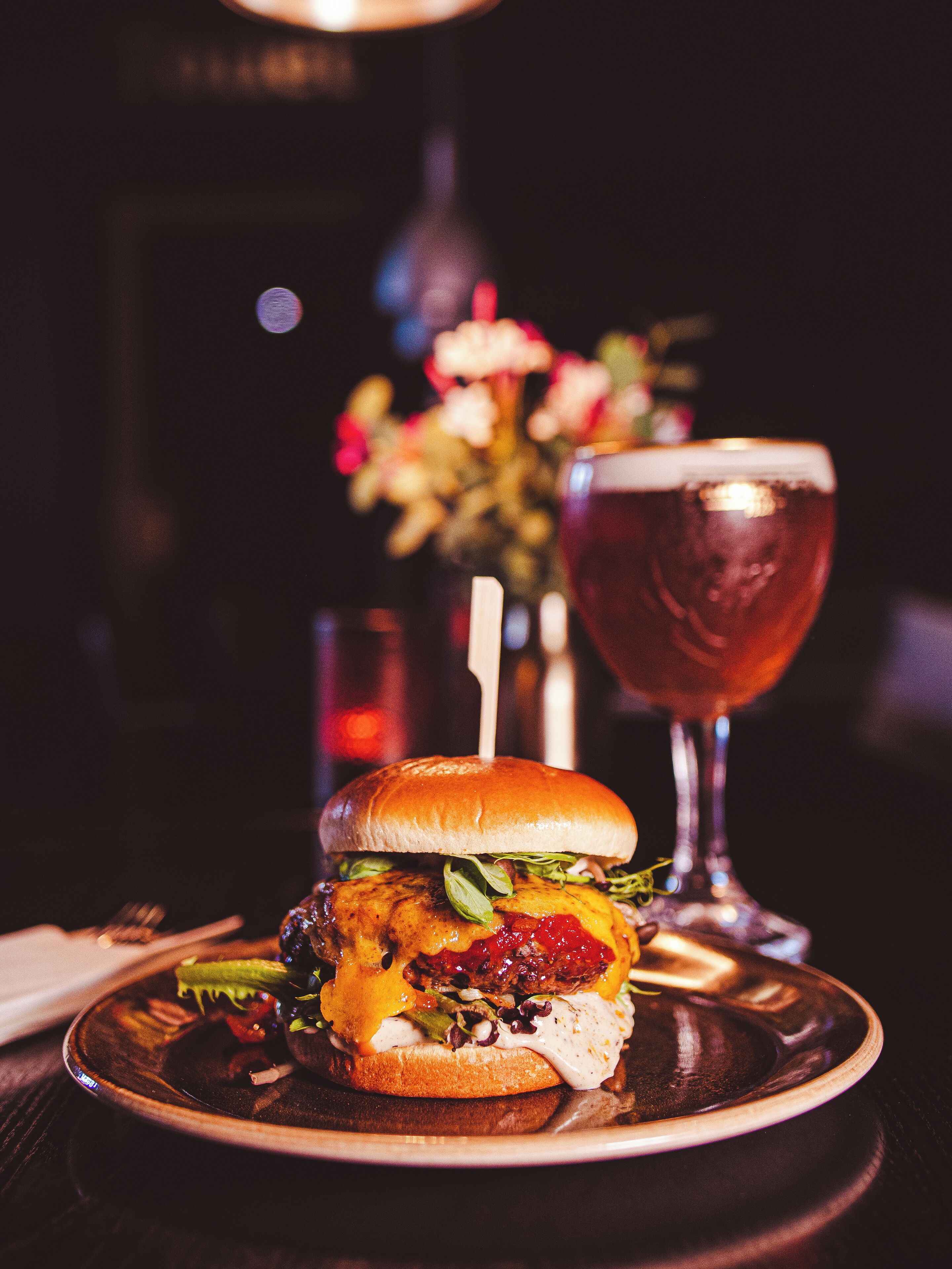 Un hamburger avec un cure-dent planté dessus, posé sur une assiette, un verre d’une boisson rouge et, au fond, un vase rempli de fleurs.
