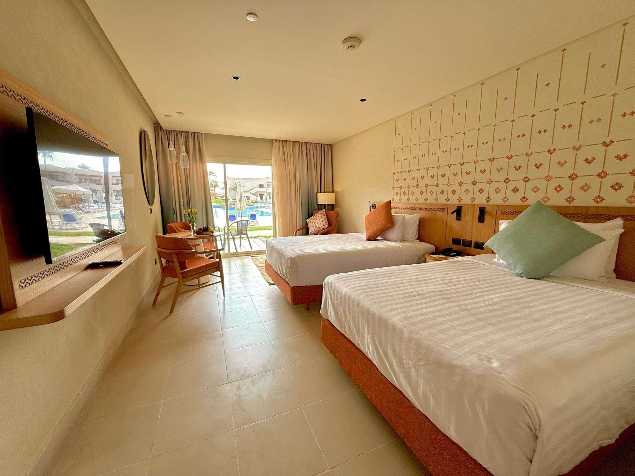 A hotel room with two beds, a TV, a desk, and a view of a swimming pool.