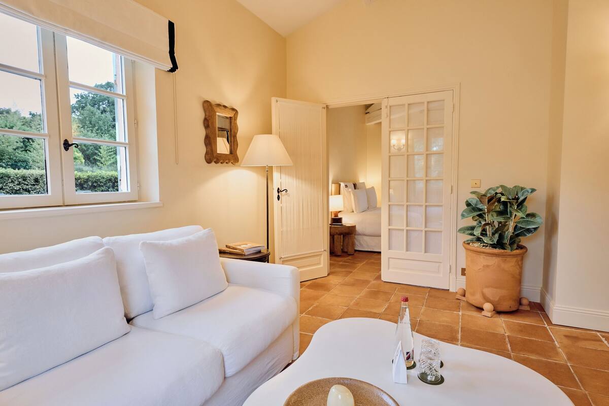 Un sofá blanco, una mesa con un florero, una lámpara, una planta en maceta y una puerta que da a otra habitación.