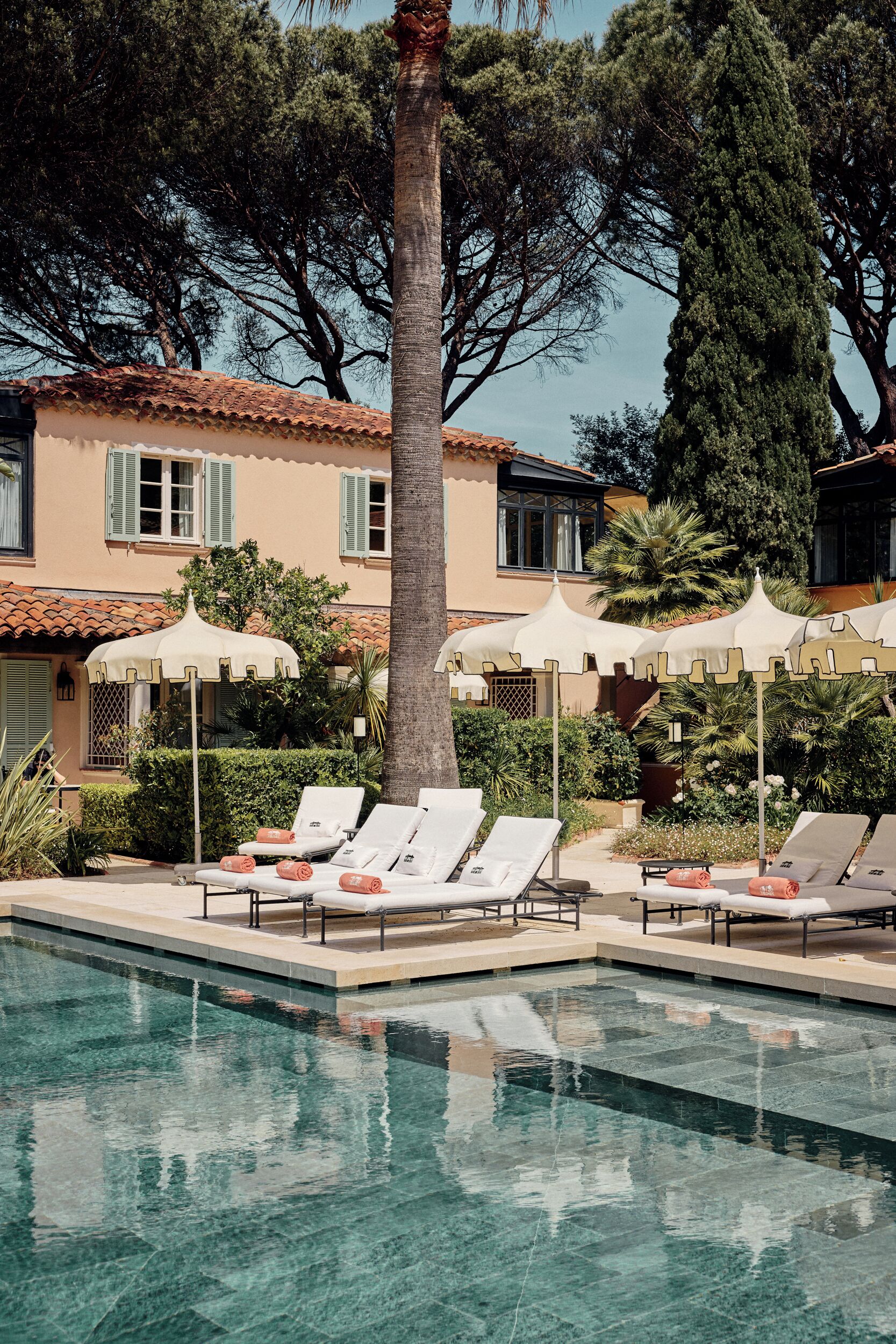 Ein Poolbereich mit Liegestühlen und Sonnenschirmen, umgeben von einem Gebäude mit einem Ziegeldach und grünen Fensterläden.