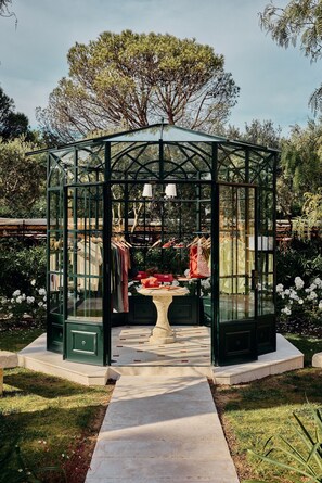 A glass greenhouse structure with a table and chairs, surrounded by a garden.