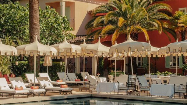 A pool area with lounge chairs and umbrellas, surrounded by palm trees and a building with a red facade. - La Bastide de Saint Tropez (Saint-Tropez)
