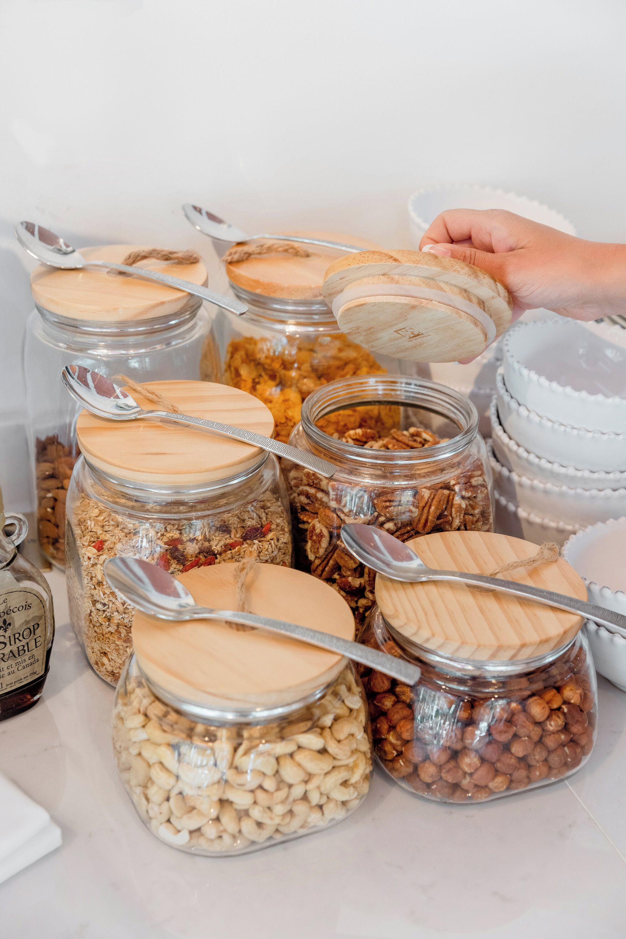 Une variété d’aliments présentés dans des bocaux en verre, fermés par des couvercles en bois, certains accompagnés de cuillères.