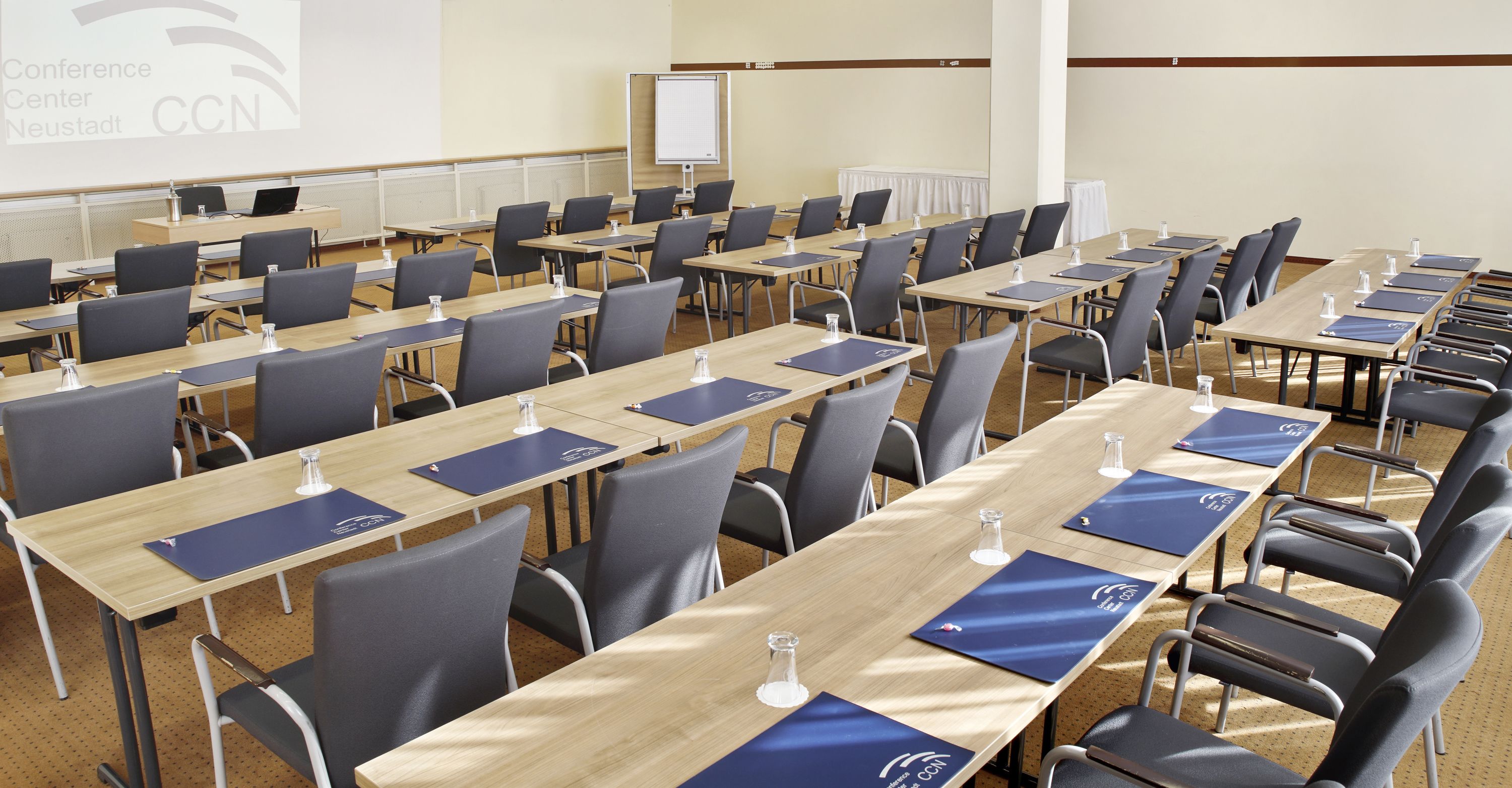 A conference room with rows of tables and chairs, a whiteboard, and a presentation screen.