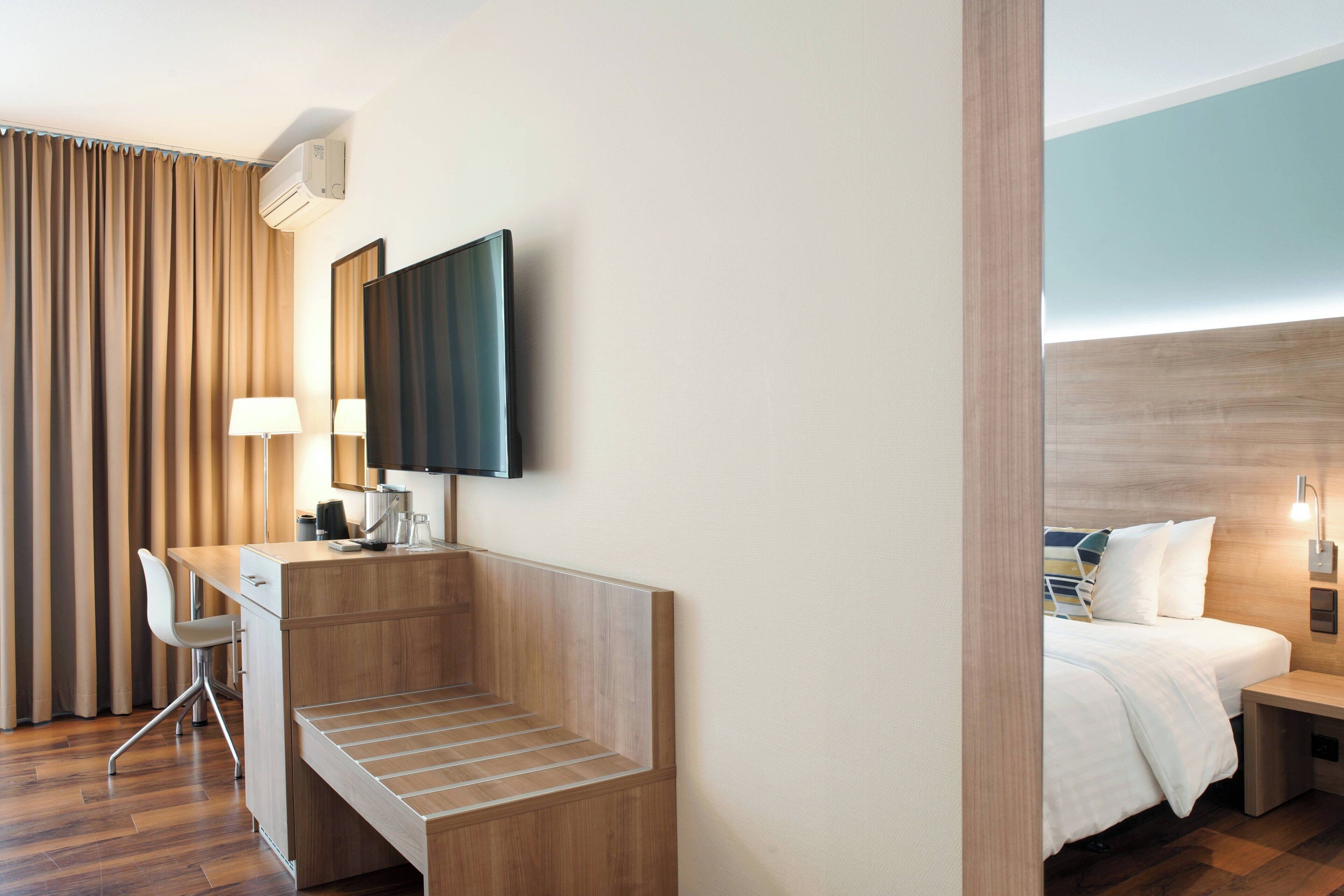 A hotel room with a wooden desk, a chair, a bed with white bedding, a TV mounted on the wall, and a window with curtains.