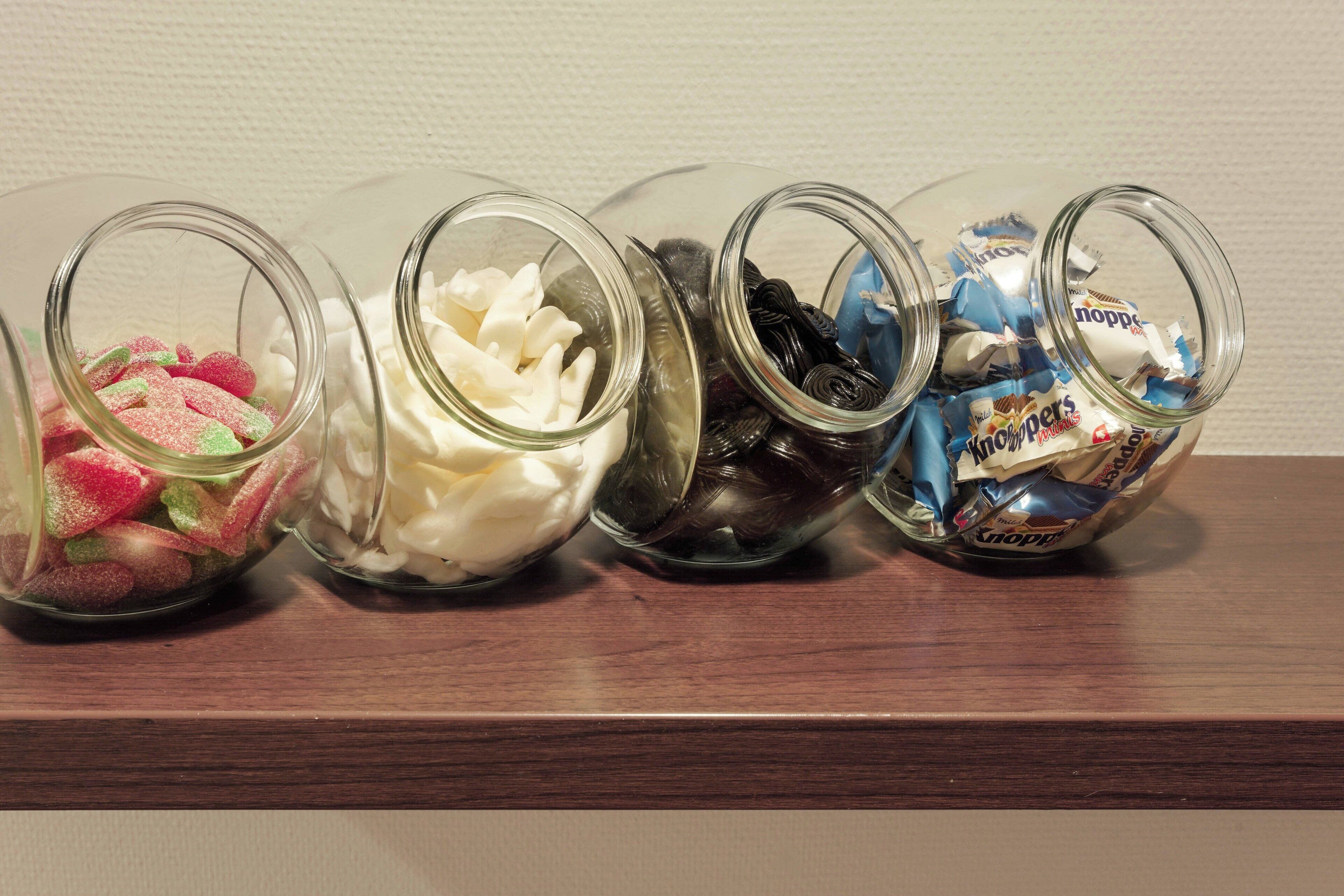 Five glass jars on a wooden shelf containing various candies.