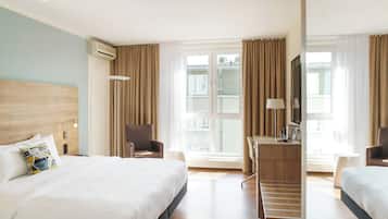 A hotel room with two beds, a desk, a chair, a TV, and large windows with curtains.