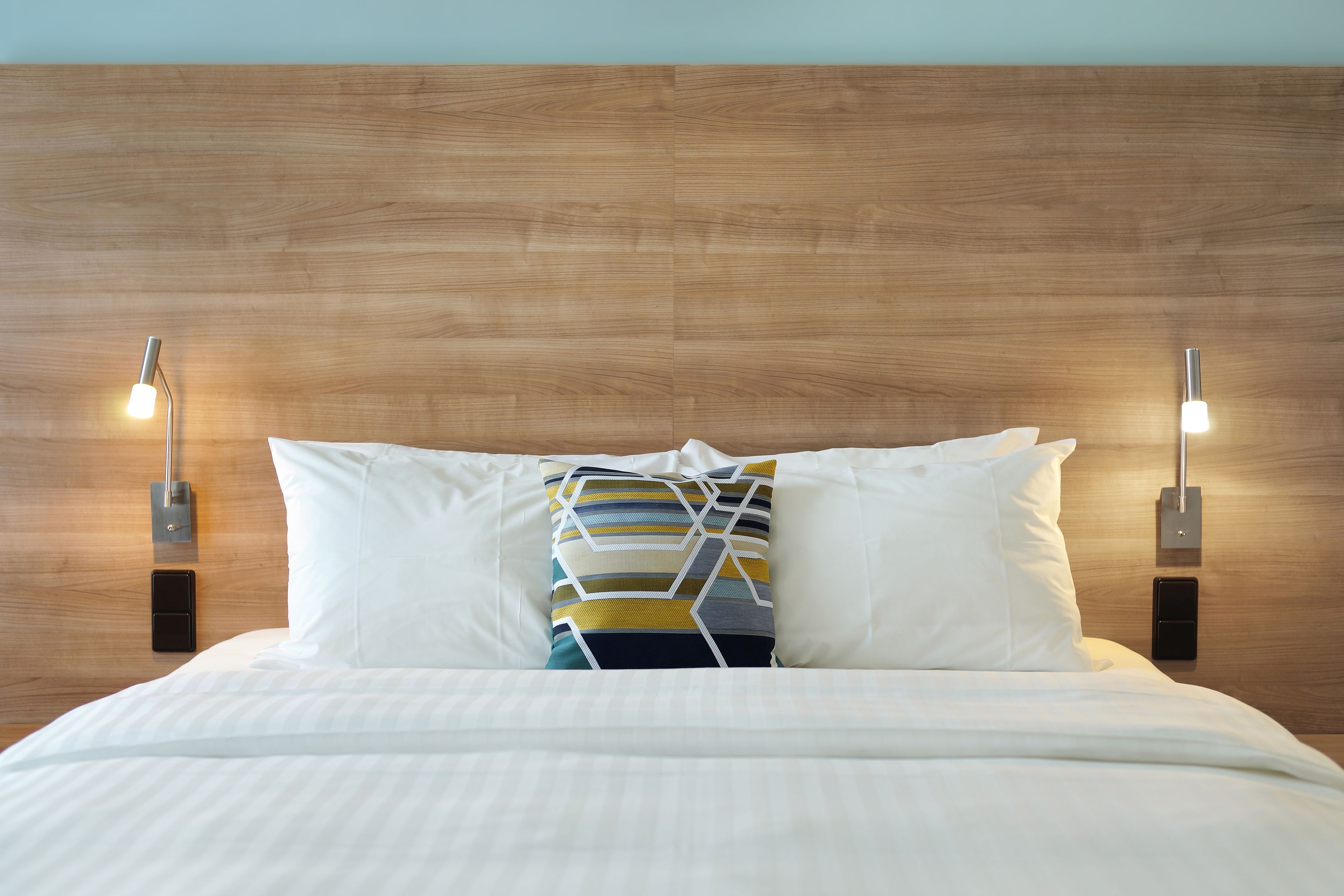 A neatly made bed with white linens and a colorful geometric pillow, flanked by two bedside lamps on a wooden headboard.