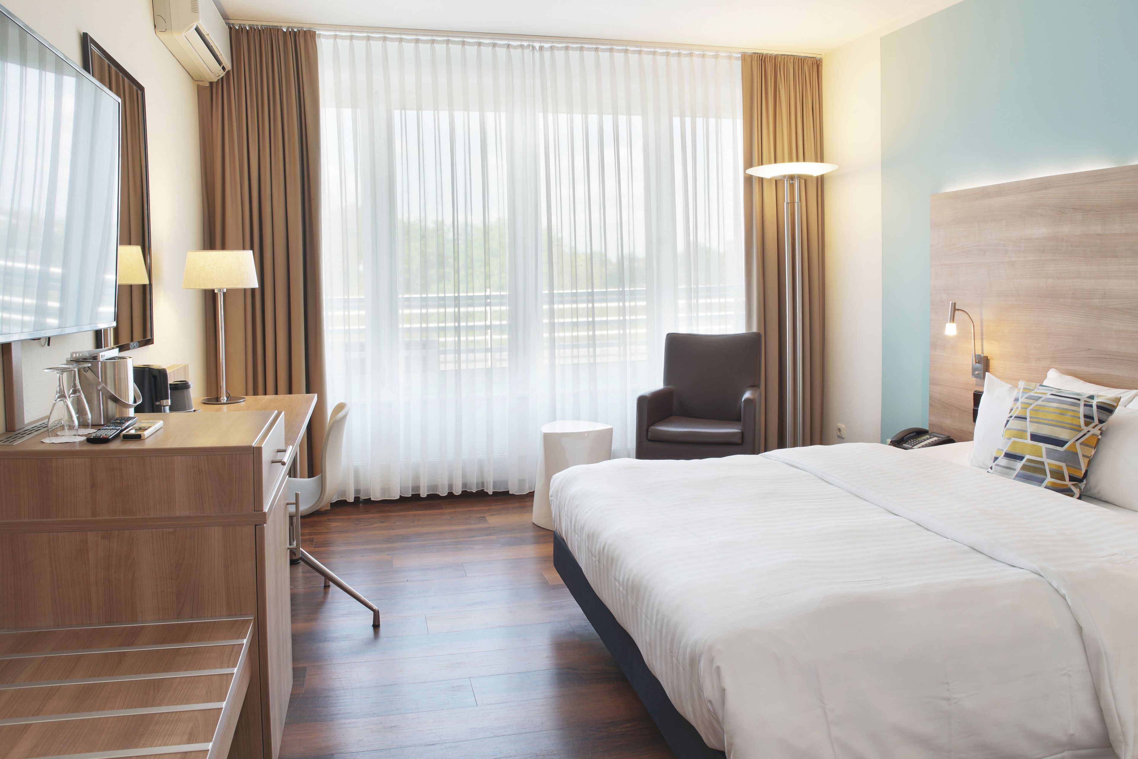 A hotel room with a bed, a desk with a computer, a chair, a lamp, and a large window with curtains.