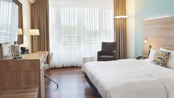 A hotel room with a bed, a desk with a computer, a chair, a lamp, and a large window with curtains.